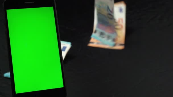 Мобильный телефон с вертикальным зеленым экраном на черном фоне, куча банкнот евро падает на черном фоне, рассчитывая деньги для будущих бизнес-инвестиций. Сохранение бумажной валюты для покупки — стоковое видео