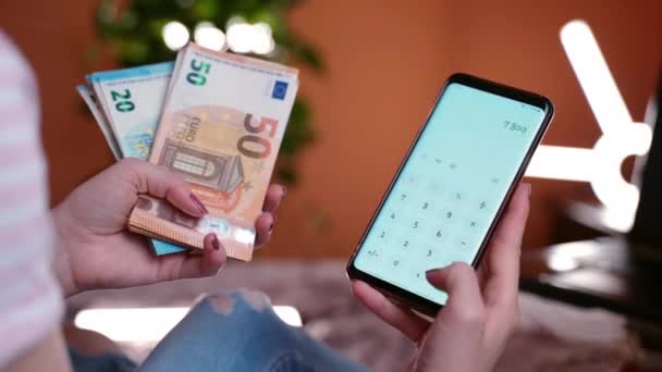 Jaarlijks financieel verslag, debet- en kredietvergelijking. Vrouwelijke accountant met een stapel bankbiljetten van 20 en 50 euro en een mobiele telefoon met rekenmachine, een vrouw die het maandelijkse inkomen van haar gezin berekent of — Stockvideo