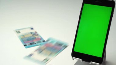 Dik yeşil ekranlı akıllı telefonun yanındaki masaya düşen euro kağıt para akışı. İş yatırımları için para kazanmak ve biriktirmek ya da yeni gayrimenkul almak. Yüksek ya da düşük gelir
