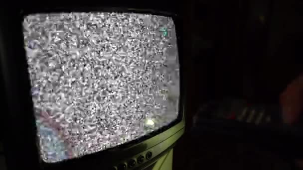 Старое ретро-ТВ транслируется в темной комнате, мужчина держит пульт дистанционного управления и переключает каналы. Устаревший пульт дистанционного управления, плохая передача сигнала, просмотр 70-х, 80-х или 90-х годов ТВ. Интерференция шума экрана — стоковое видео