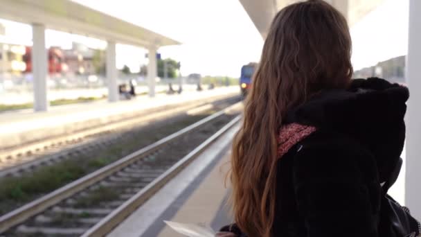 Linda menina de cabelo vermelho com lábios vermelhos em pé na plataforma da estação com bilhetes e sorrindo na câmera enquanto o trem chega. Passageiro do sexo feminino à espera de viajante público na estação urbana, viagem de — Vídeo de Stock