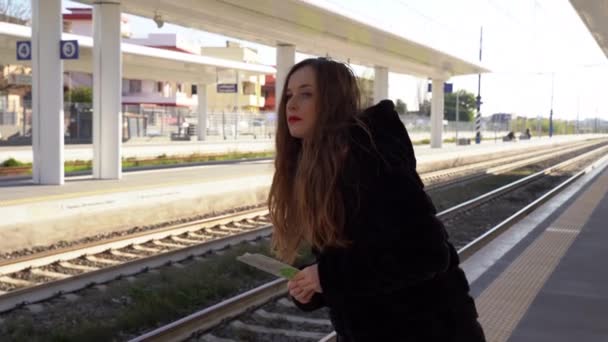 Fiatal vörös hajú lány piros ajkakkal áll az állomás peronján, és várja a vonatot, lány kezében utazási jegyek mindkét irányban. Arra gondolok, hogy a női utas egyedül várakozik a peronon. — Stock videók