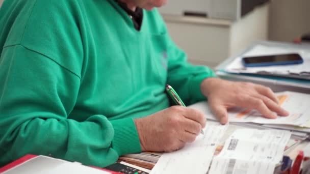 Praca w domu w kwarantannie koronawirusowej, papierkowa robota w oddali. Starszy mężczyzna pracujący przy stole w apartamentowcu, stary lekarz zapisujący coś długopisem na kartce papieru, czytający i notujący — Wideo stockowe
