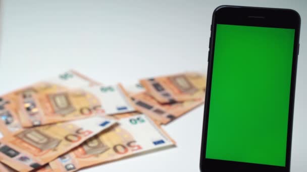 Smartphone avec écran vert vertical chroma clé et la chute des factures en euros sur fond. Un paquet de papier-monnaie à table, grand prix de loterie en billets de 20 et 50 euros. Concept de jackpot de trésorerie — Video