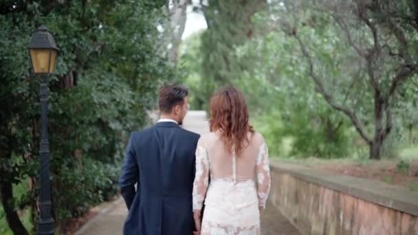 짙은 청색 양복을 입은 신랑 과아름다운 레이스 달린 긴 우유 색깔의 옷을 입은 젊은 부부의 훌륭 한 의상이다. 놀라운 녹색 공원을 걷고 있는 행복 한 신혼 부부와 — 비디오