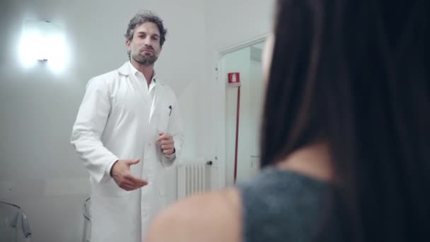 Handsome male doctor in white lab coat meeting young female patient on reception in medical clinic. Friendly physician talking to woman and inviting her in room for further diagnostic consultation — Stock Video
