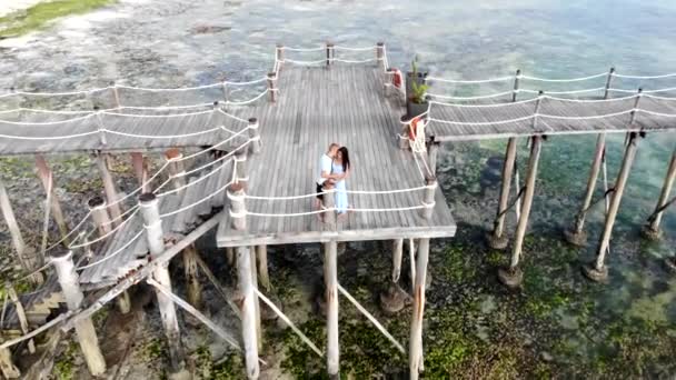 Feliz pareja casada pasando su luna de miel en el increíble complejo vacacional de Zanzíbar. Vista aérea de dos enamorados que se quedan en un puente de madera entre el mar, abrazándose y besándose — Vídeo de stock