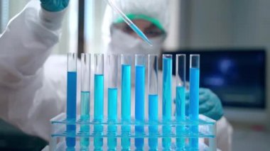 Koruyucu kıyafetli konsantre laboratuvar araştırmacısı hareket eden mikrop videosuyla monitörün arka planında oturuyor ve steril test tüplerine mavi reaktör bırakıyor. Profesyonel virüs uzmanı.