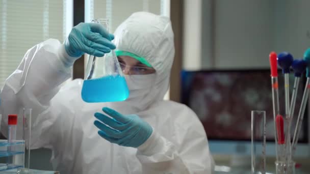 Laboratorní vědec v ochranném obleku, masce a brýlích pracující s nebezpečným chemickým roztokem ve výzkumné laboratoři, chemička používající sterilní pipetu a odměřovací baňka na výrobu vakcíny proti — Stock video
