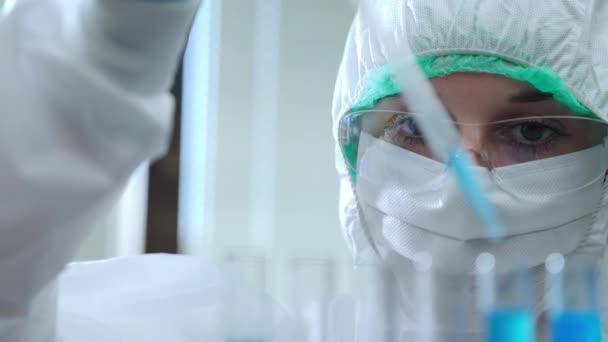 Close-up van vrouwelijke laboratoriumonderzoeker in beschermend pak en bril met steriele meetpipet en druppeloplossing in reageerbuizen. Geconcentreerde chemicus die medische experimenten uitvoert in het laboratorium — Stockvideo