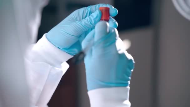 Κοντινό πλάνο των χεριών του μικροβιολόγου με προστατευτικά μπλε γάντια που συγκρατούν το σωλήνα εξέτασης αίματος και καταγράφουν τα δεδομένα της ανάλυσης. Ιατρικό εργαστήριο γιατρός ανάλυση πλάσματος αίματος του μολυσμένου ασθενούς, coronavirus — Αρχείο Βίντεο