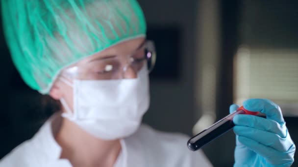 Специалистка-медик, работающая в лаборатории с плазмой крови, врач в защитных перчатках, маске и очках, держащая в руках трубку для анализа крови и записывая на ней данные анализа — стоковое видео
