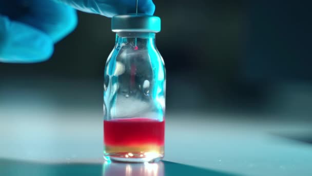 Красный химический раствор выпадает из стерильного шприца внутри прозрачного флакона с желтой жидкостью. Крупный план рук врачей в стерильных голубых перчатках для дальнейшего медицинского анализа — стоковое видео