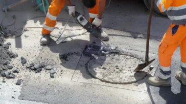 Koruyucu turuncu üniformalı iki işçi yolda beton kırmak için ağır matkap kullanıyor, caddede tıkanmış kanalizasyon açmak için inşaat alanında sıkı çalışıyor.
