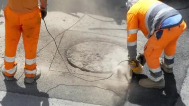 İnşaat alanında çalışan koruyucu üniformalı adamlar yolda çimento kesmek için elektrikli matkap kullanıyorlar. Kanalizasyon kapağını açmak için beton kıran inşaat işçisi.