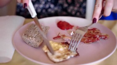 Kadınların elleri leziz kahvaltı, kızarmış yumurta, pastırma dilimleri ve pembe tabakta domates soslu kızarmış çavdar ekmeği kesiyor ve yiyor. Sağlıksız yemek, Amerikan kahvaltısı.