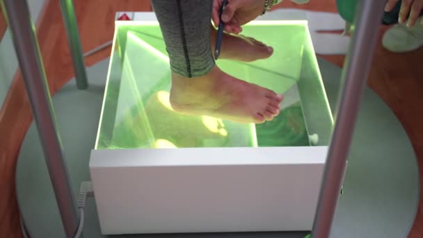 Çıplak ayaklı hasta saydam cam yüzeyde yeşil neon ışıkla kalırken erkek ortopedist doktor ayak ve kemik durumlarını inceleyip kontrol ediyor. Düz taban ve sağlık sorunları — Stok video