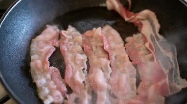 Aşçı tavada yağlı pastırma dilimleri pişiriyor, tava yüzeyinde şerit çevirmek için ahşap çatal kullanıyor. Kıtır kıtır dilimler cızırdıyor ve sigara içiyor, lezzetli ve besleyici bir kahvaltı hazırlıyor.