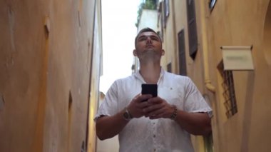 Genç yakışıklı adam, yüksek binalarla antik dar caddelerde yavaş yavaş yürüyor, akıllı telefon ve online GPS haritalarıyla doğru yöne dönüyor. Beyaz adam binaya bakıyor.
