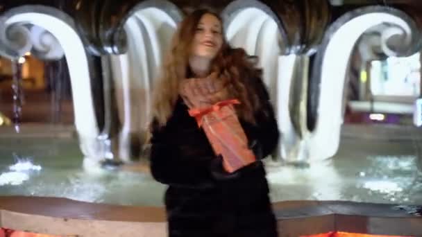 Glücklich tanzende Frau mit Weihnachtsgeschenk in der Hand auf der beleuchteten Straße, lächelnde Frau genießt Weihnachtsgeschenk und zeigt aufrichtige Emotionen vor der Kamera. Konzept der Weihnachtsfeier — Stockvideo