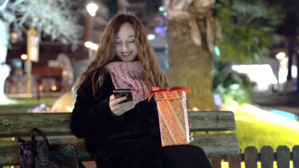 Mujer bonita sentada en el banco en el parque, la celebración de la Navidad presente en las manos y el uso de teléfonos inteligentes, sonriente mujer charlando en línea con amigos, el envío de mensajes de Navidad a los miembros de la familia y — Vídeo de stock