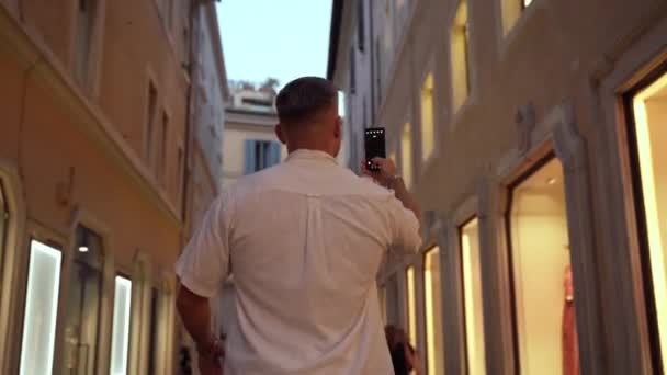 Vista trasera del hombre en traje casual caminando por la antigua calle con boutiques de lujo iluminadas y tomando fotos en el teléfono inteligente. Joven turista tomando fotos en el teléfono móvil para la memoria, hombre — Vídeo de stock