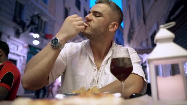 Handsome man in casual outfit sitting outdoors in a pub and eating delicious italian pizza and drinking draft beer, young man in a cafe on amazing building exterior background and crowded evening — Stock Video