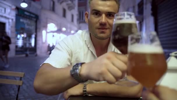 Jovem homem bonito sentado ao ar livre em um café, batendo copos com amigo e beber cerveja rascunho fresco, homem sorrindo reunião com amigo depois de muito tempo, falando e degustando cerveja em um pub — Vídeo de Stock