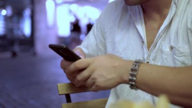Erkeğin elleri cep telefonu tutarken ve şehrin aydınlanmış arka planında mesajlar yazarken, adam dışarıda kafede oturuyor ve akıllı telefon kullanıyor, internette geziniyor veya iletişim kuruyor.