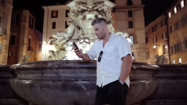 Concentrated man standing on amazing Roman building exterior background and leaning on ancient dark marble fountain with illuminated statues and flowing water. Serious man using smartphone, chatting — Stock Video