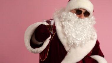 Modern Noel Baba 'nın kırmızı geleneksel Noel elbisesi ve siyah gözlükleriyle elinde bir bardak alkollü içecek tutarken ve kamerada gösterirken orta boy fotoğrafı. Sarhoş Noel Baba 'nın dinlenme zamanı.