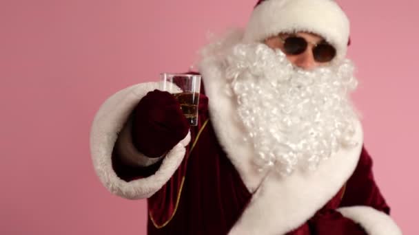 Średnie ujęcie współczesnego Mikołaja w tradycyjnym czerwonym garniturze świątecznym i czarnych okularach trzymających szklankę napoju alkoholowego i pokazujących przed kamerą. Czas odpoczynku pijanego Mikołaja na przyjęciu, Boże Narodzenie — Wideo stockowe