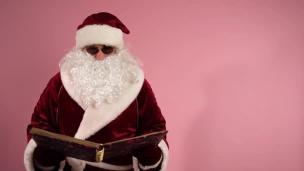 Bärtiger älterer Herr im Nikolauskostüm und mit dunkler Brille, der ein Weihnachtsbuch in der Hand hält, liest und den Menschen frohe Weihnachten und einen guten Rutsch ins neue Jahr wünscht. Konzept für Weihnachtsferien — Stockvideo