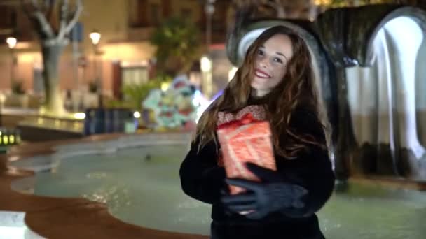 Retrato de mujer bonita bailando con regalo de Navidad en las manos sobre fondo de fuente de agua fluida iluminada. Decoraciones de Navidad en la calle, estado de ánimo de celebración — Vídeo de stock