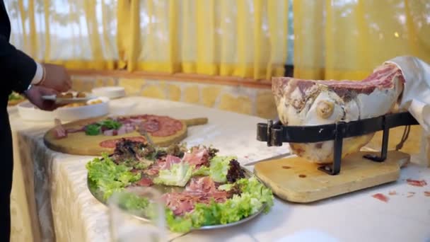 Mesa buffet bellamente decorada en un restaurante de lujo u hotel, huésped en traje que sirve a sí mismo con deliciosa cocina italiana. Famosas rebanadas de jamón italiano en el plato de madera, los huéspedes degustación de carne — Vídeos de Stock