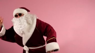 Kırmızı geleneksel kostümlü kaygısız Noel Baba ve pembe arka planda dans eden koyu gözlükler ve kamera önünde el kol hareketleri yapan. Komik, modern Noel Baba kutlamada Noel performansı sergiliyor.