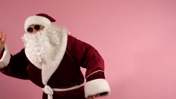 Carefree Santa Claus in red traditional costume and dark glasses dancing on pink background and gesticulating with hands on camera. Funny modern Santa showing christmas performance at the celebration — Stock Video