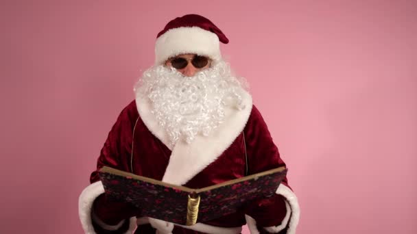 Serious Santa Claus en traje rojo y gafas oscuras de pie sobre fondo rosa y sosteniendo el libro mágico de Navidad en las manos, barbudo libro de lectura de Santa con placer. Santa Clause relajarse en la habitación y — Vídeos de Stock