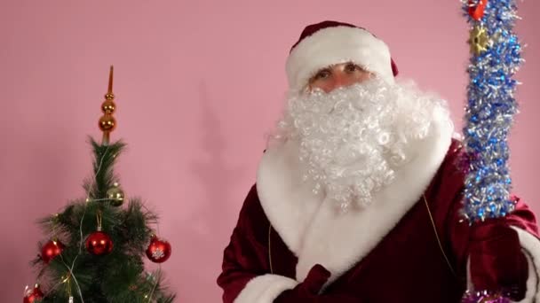 装飾されたクリスマスツリーの近くのピンクの背景に立っている赤いクリスマスの衣装でサンタクロースは、装飾された棒を保持し、手袋で手でジェスチャーをカメラに話しています。髭を生やしたサンタが人々を誘う — ストック動画