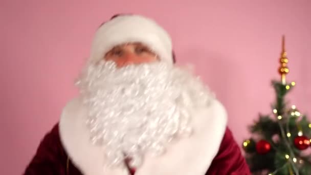 Mittlere Aufnahme des lustigen Weihnachtsmannes, der ein Glas Cognac in der Hand hält und vor der Kamera tanzt, des Weihnachtsmannes im traditionellen roten Anzug, der auf rosa Hintergrund tanzt und den Weihnachtsbaum leuchten lässt. Betrunkener Weihnachtsmann genießt Party, singt — Stockvideo