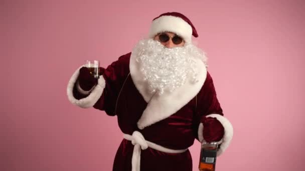 Grappig dronken Kerstman in bril met glas met cognac in de ene hand en fles met alcohol in de andere hand, vrolijke Kerstman in rood traditioneel kostuum genieten van ontspannen tijd op kerstfeest — Stockvideo