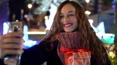 Uzun kıvırcık saçlı güzel bir kadın akşam parkında bankta oturuyor, elinde Noel hediyesi tutuyor ve ailesiyle ya da arkadaşlarıyla internetten video görüşmesi yapıyor. Güzel bir kadın insanlarla konuşuyor.