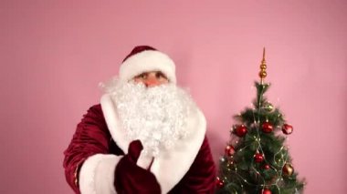 Geleneksel kırmızı kostümlü neşeli Noel Baba kamerada dans ediyor, başparmak işareti gösteriyor ve elinde farklı hediyelerle büyük kırmızı bir Noel çantası tutuyor. Noel Baba pembe arka planda dans ediyor ve süslü Noel ağacı