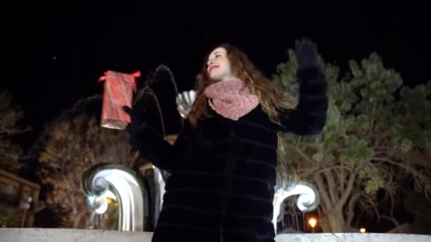 Vacaciones de Navidad y estado de ánimo atmosférico feliz alrededor, mujer sonriente en ropa de invierno bailando en el parque por la noche en el fondo de la fuente de agua que fluye y la celebración de Navidad presente en las manos — Vídeo de stock