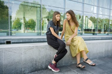 İki kız kardeş caddede binanın dışında karşılaştı. Oturup konuşuyorlar. Şehir, yaz, güneşli gün.