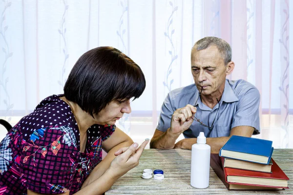 A woman shows a man eye lenses. A man is sitting at a table in a room. Carefully look. He holds glasses in his hands.