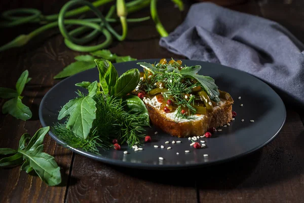 Toast with curd cheese, fried garlic arrows and herbs on a dark background.