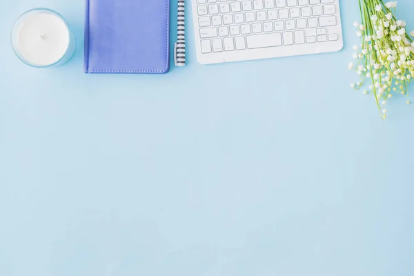 Flat lay blogger or freelancer workspace with a notebook, keyboard and lily of the valley flowers on a blue background