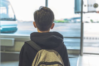 Sırt çantası olan bir çocuk, havaalanındaki cam kapının yanında durup uçağa giden otobüse bindirmeyi bekliyor. Gidiş salonu