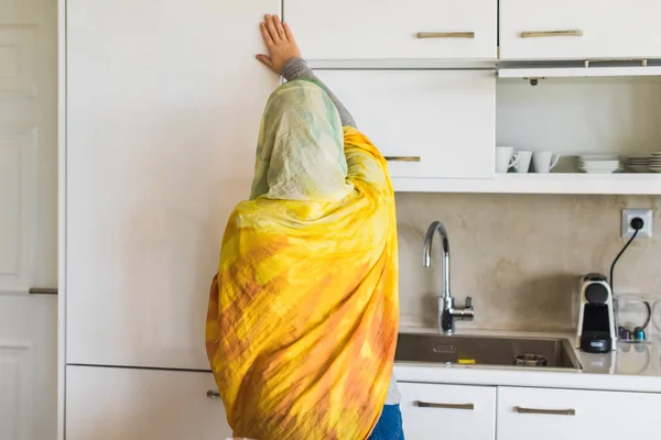 Back view on a muslim woman in a scarf opening door of a fridge on the kitchen. Preparation of breakfast